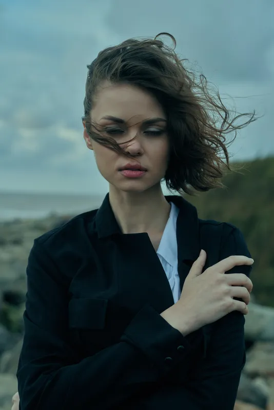 Moody outdoor image of a woman with windblown hair near the coast, wearing a dark jacket, thoughtful expression and wind catching fabric