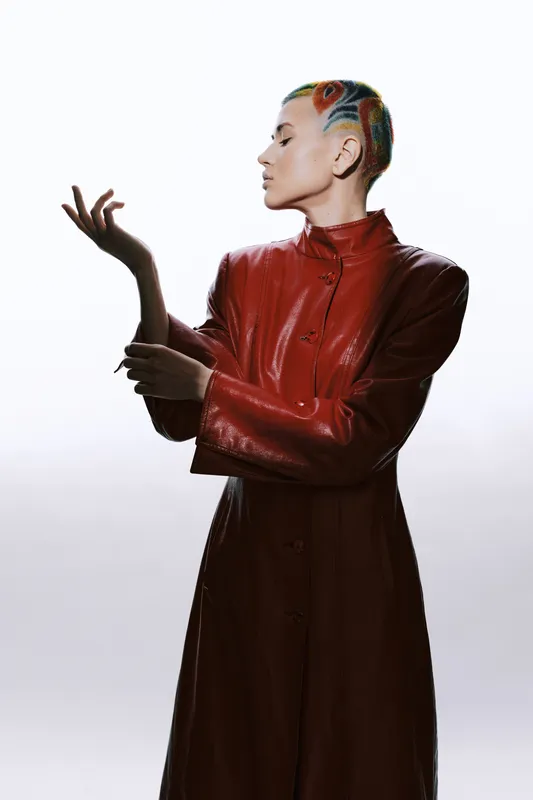 woman with colorful braided hair wearing a red leather coat poses elegantly against a bright white background, showcasing fashion and style in a modern portrait.