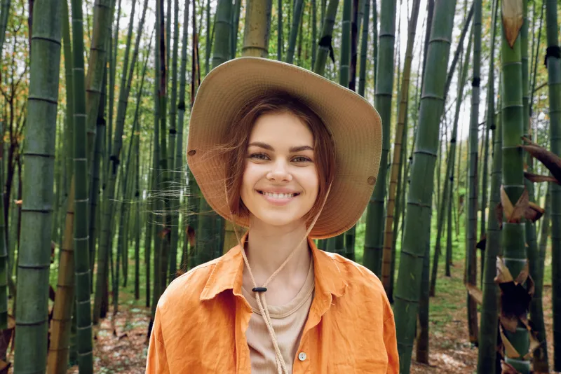 Woman portrait wearing a wide brim hat and orange shirt in a bamboo forest outdoors, nature background. Casual travel scene with a warm smile, sunlight filtering through tall green stalks.