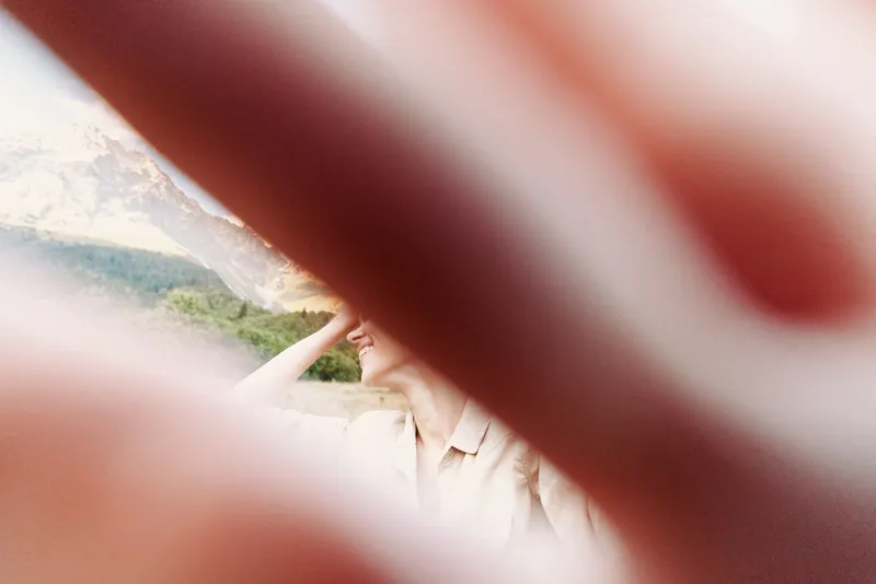 A candid outdoor portrait of a woman in a light jacket, seen through a blurred foreground, capturing a spontaneous lifestyle moment of curiosity and calm.