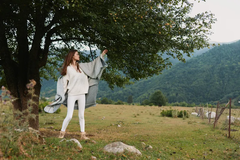 Young woman outdoors in a grassy field lifts a light shawl against the breeze, smiling softly with mountains and trees behind her, wearing a white sweater for a casual lifestyle moment.