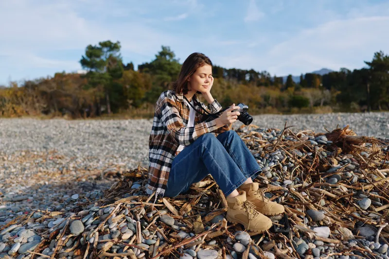 Woman photographer sits on a rocky shoreline with a camera, wearing plaid shirt and boots, capturing coastal textures and natural light in an outdoor scene