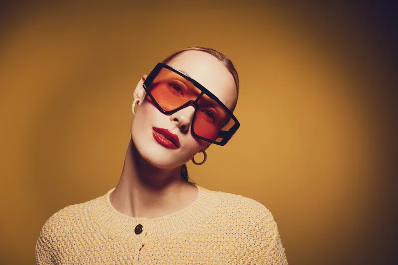 Stylish woman with slicked back hair wears large orange sunglasses and hoop earrings. She poses confidently against a warm yellow background in a knitted sweater. Fashion concept.