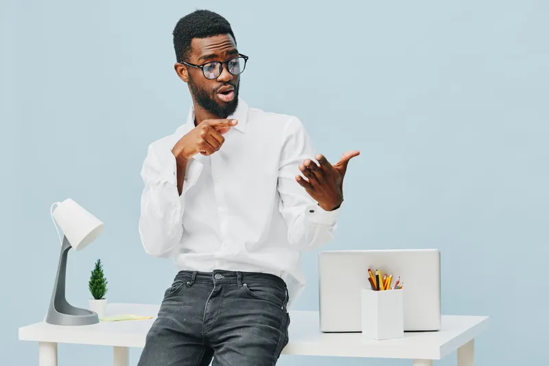 Confused man with glasses and beard wearing white shirt, standing at modern desk with laptop and stationery, gesturing with hand, looking puzzled, blue background, studio shot. People lifestyle concep