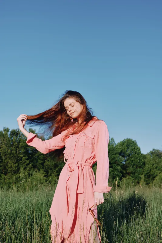 Young woman in a flowing pink dress enjoying a sunny day outdoors, showcasing beauty and freedom in nature, with vibrant greenery and a clear blue sky