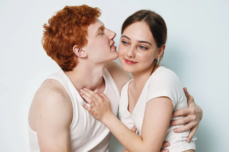 Couple, woman and man embracing in white clothing with affection and closeness. Intimate, young adult pair showing love, care and connection in studio with smiles and gentle touch.