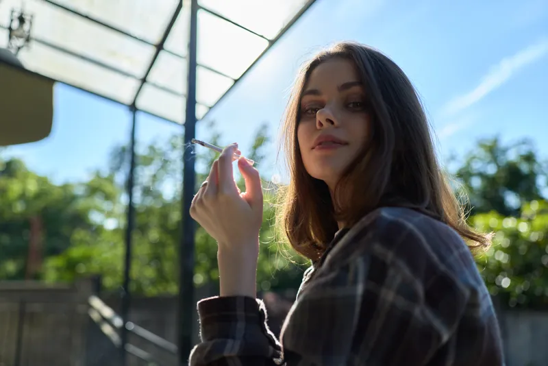 Casual outdoor portrait of a woman in plaid shirt smoking a cigarette, relaxed lifestyle, sunny day, natural light, urban garden backdrop, candid and calm