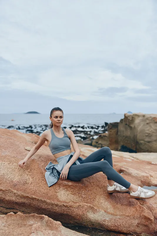 Stylish young woman in sportswear sitting on rocks by the beach, showcasing active lifestyle, fitness emotions, and coastal serenity in overcast weather