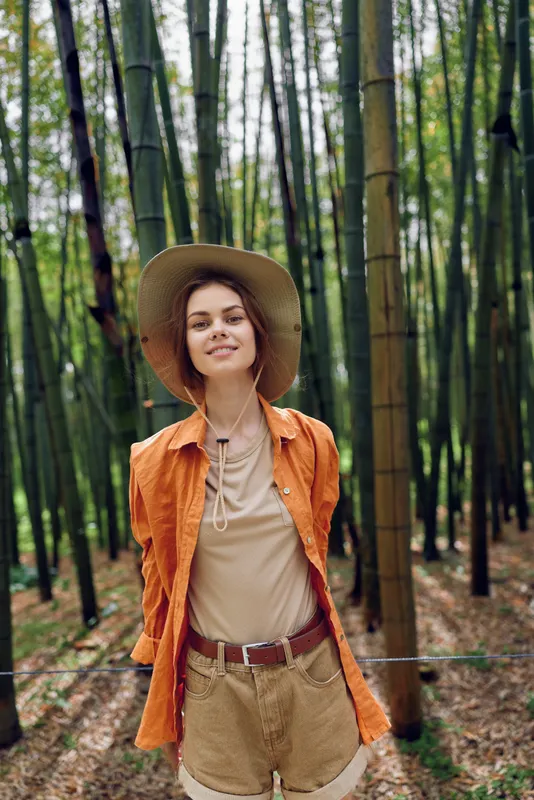 Woman in bamboo forest wearing straw hat and casual orange shirt, smiling portrait amid tall green stalks, nature walk in summer outfit, relaxed pose and soft natural light.