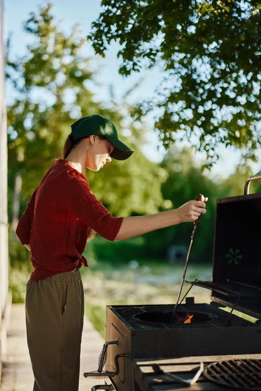 Young woman grilling outdoors in a sunny garden, wearing casual clothing and a cap, enjoying a joyful summer moment with a barbecue