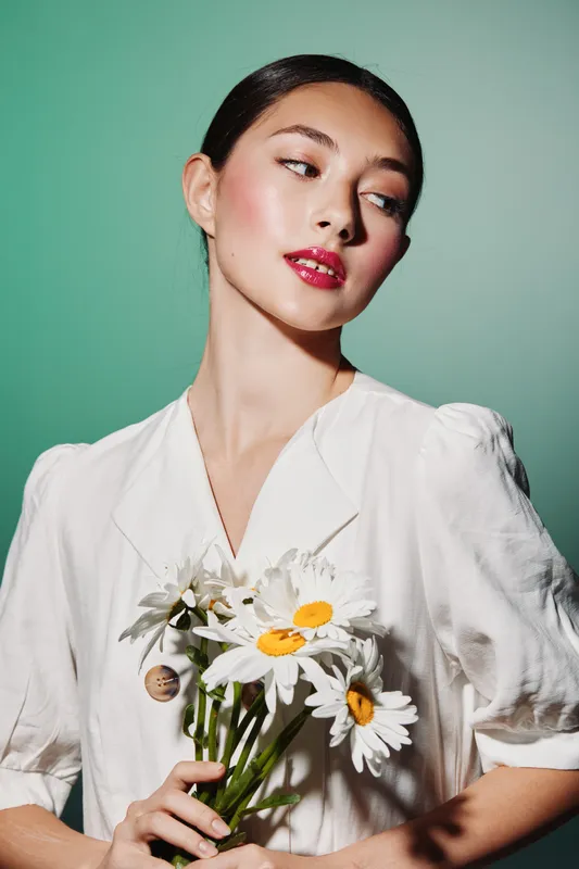 Young woman with bright makeup and glossy red lips poses holding daisies wearing a white vintage-style blouse on green background studio shot. Beauty fashion creative.