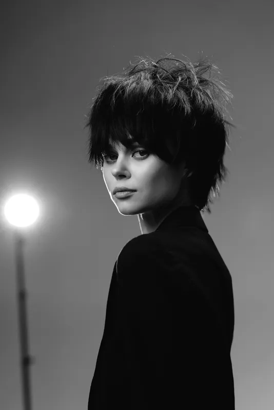Black and white portrait of a young woman with short hair and dark clothing in moody lighting.