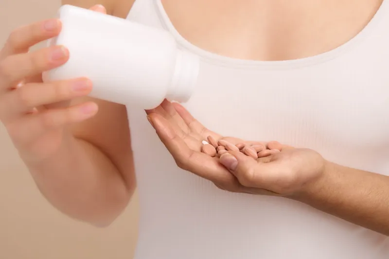 Asian woman pours supplement pills from a bottle into her hand in a closeup, soft studio lighting. The scene conveys focus on health, care, and personal wellness.