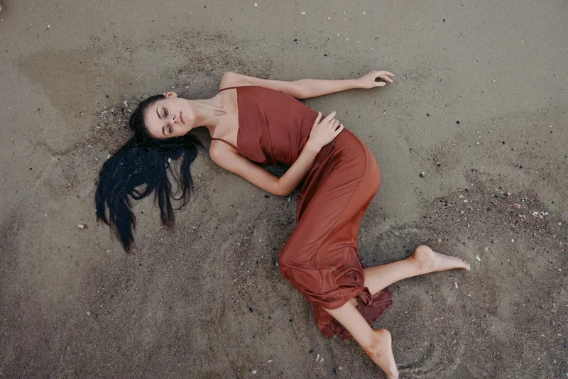calm woman lying on sandy beach wearing a red dress, relaxed and peaceful with long black hair spread out, natural and serene mood