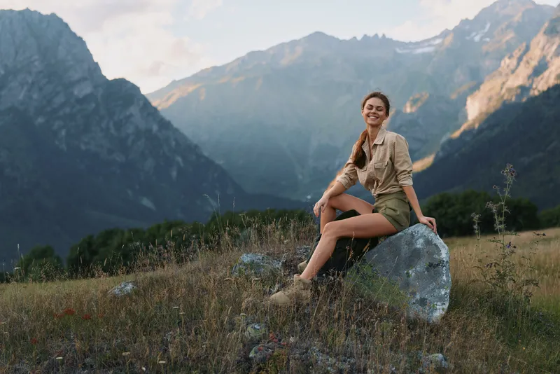 A young woman sits atop a rock in a mountain meadow, smiling softly as sunlight bathes the alpine landscape, conveying a moment of peaceful adventure outdoors.