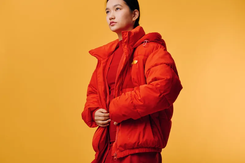 Young woman in orange winter jacket stands against yellow background with hands adjusting zipper, wearing warm clothing for cold weather, casual style and thoughtful expression.