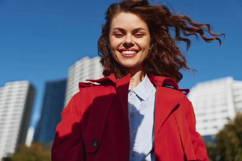 Young woman with curly hair smiles joyfully wearing a red coat and white shirt outdoors in an urban setting on a clear sunny day.