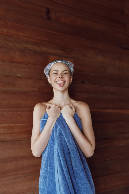 Young woman wrapped in a blue towel with a shower cap smiling playfully against a wooden background indoors, showing joy and freshness after a bath or spa.