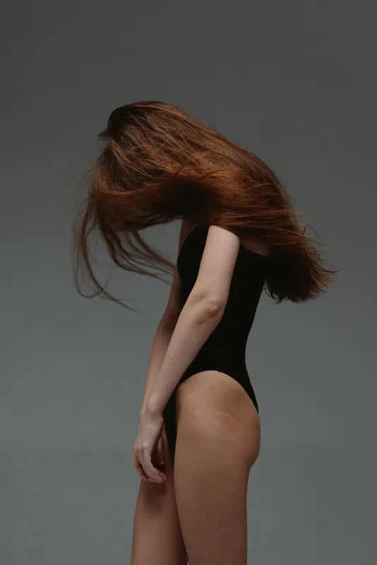 Woman with long red hair flipping it back in black bodysuit against gray background