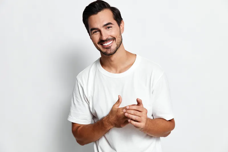 Happy smiling young man in a casual white t-shirt looking at the camera, showcasing a cheerful expression with hands clasped together against a plain background.
