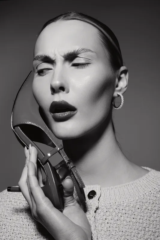 Close-up of a young white woman holding a shoe to her face with an expressive confused look in black and white studio lighting.