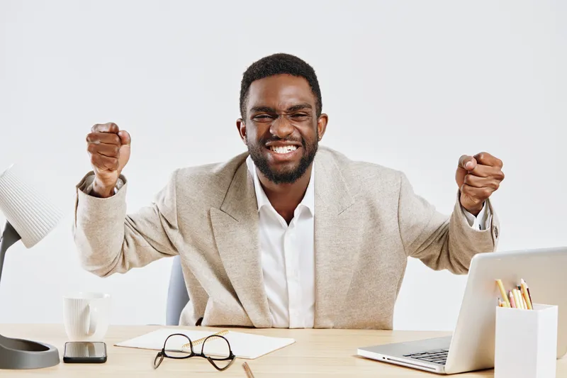 Smiling young African American man in beige blazer celebrating success at work, sitting at desk with laptop, glasses, and coffee mug, enthusiastic gesture, positive emotions, professional office envir