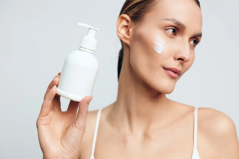 A young woman with clear skin holds a white lotion bottle while applying cream on her cheek in a bright studio. Concept for skincare, beauty routine, and facial care products.