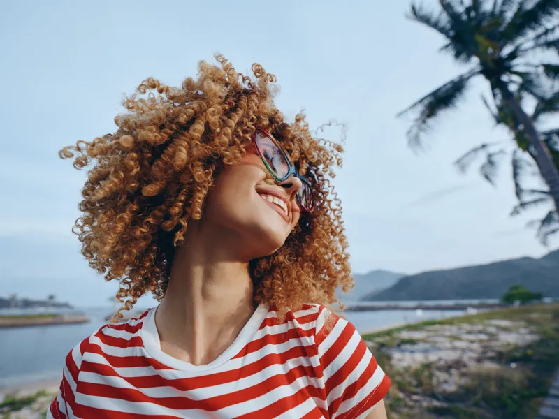 Vibrant portrait of a woman with curly hair and sunglasses, wearing a red and white striped shirt, against a coastal backdrop of water, palm trees, and bright sky.