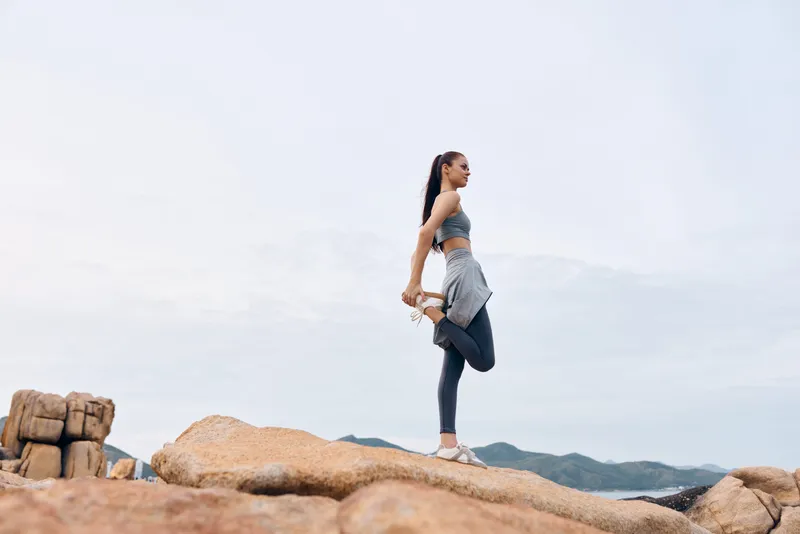 Young athletic woman stretching on rocky beach, embodying fitness and serenity in activewear with a calming natural background and soft colors