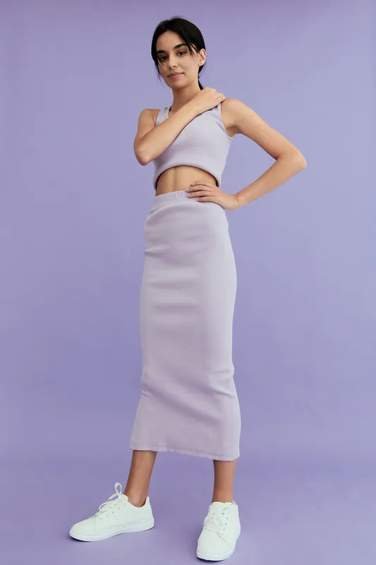 young woman wearing lilac ribbed crop top and matching long skirt posing confidently against solid purple background with white sneakers, casual fashion style
