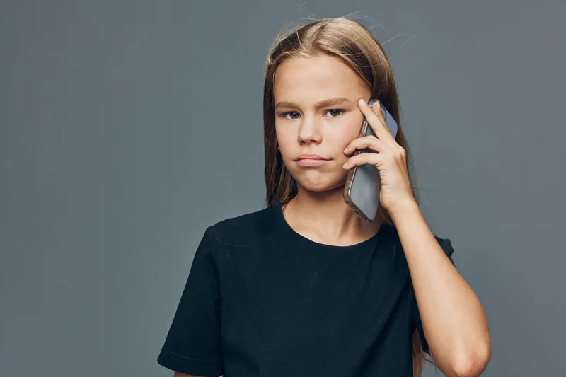 Girl with serious expression talking on phone against gray background, wearing black t shirt. Communication, technology and teenager using smartphone for conversation or connection.