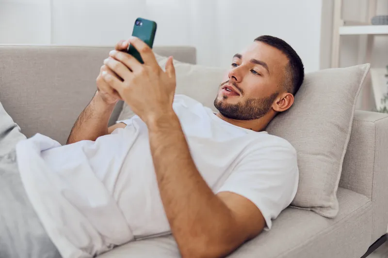Relaxed man using a smartphone while lying on a cozy couch, showcasing modern lifestyle and comfort in a bright, minimalistic room with soft colors.