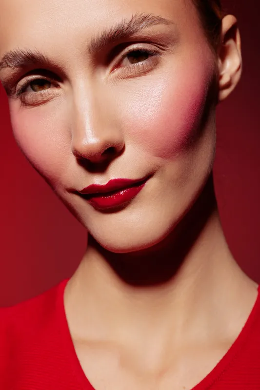 Close-up portrait of a woman with red lipstick and blush on a red background. The image highlights makeup, beauty, and fashion concepts with soft lighting and smooth skin texture.