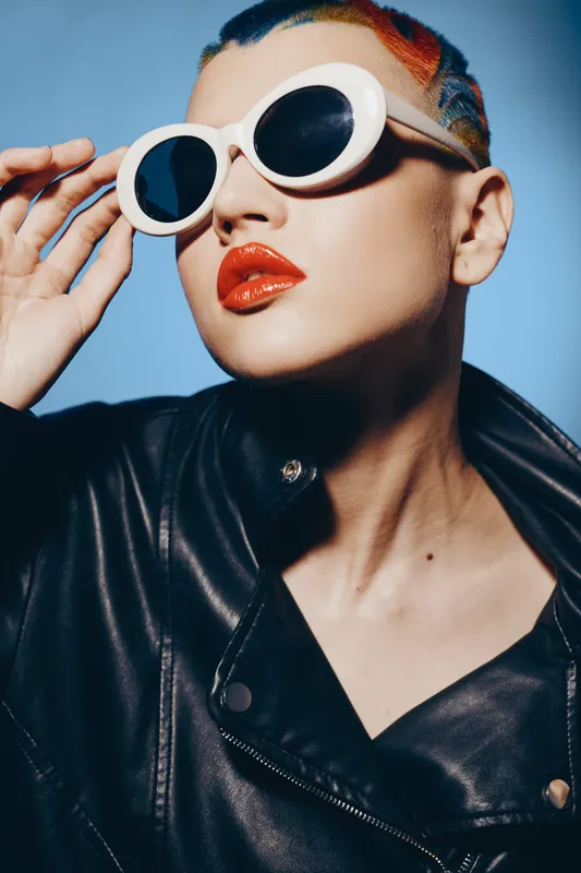 Woman with colorful shaved hair wearing oversized white sunglasses and glossy red lipstick, dressed in a black leather jacket against a blue background.
