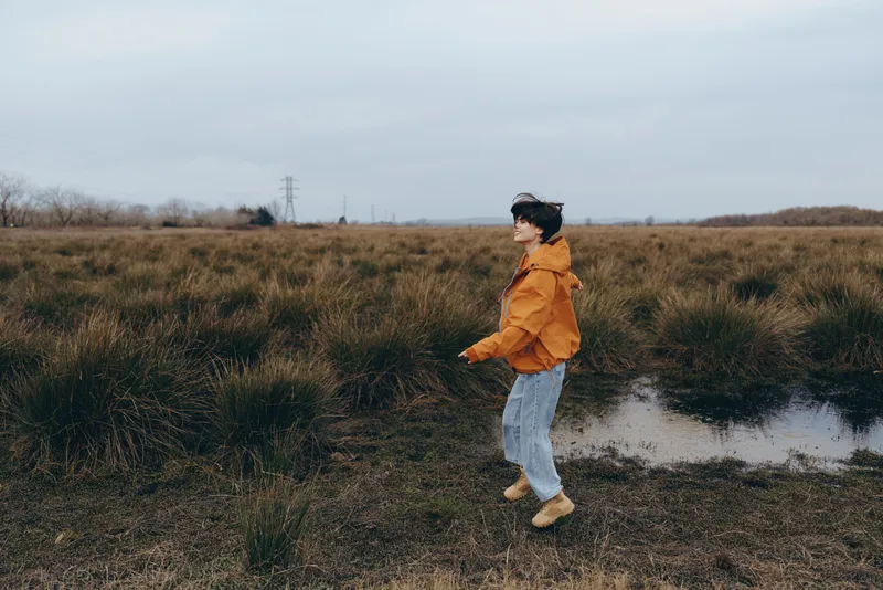Woman lifestyle outdoors walking in grassy field beside stream, cloudy sky, yellow jacket, jeans, hat, casual stride, side view, spring nature, calm mood, candid moment, near trail edge