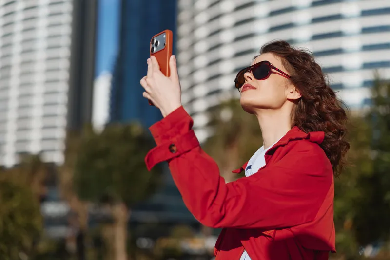 Young woman in red coat and sunglasses taking selfie with smartphone outdoors, urban background, sunny day, confident expression, casual style