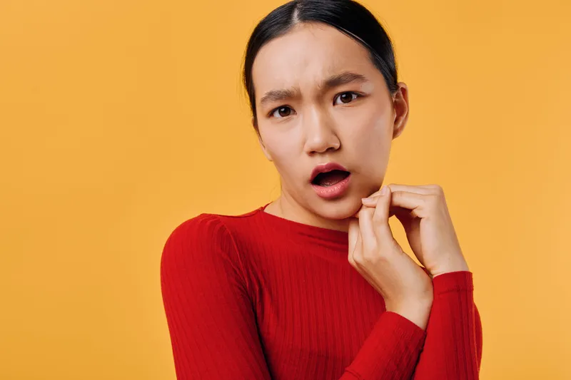 Young woman in red sweater with worried expression touching chin, standing against orange background. Concerned female person looks surprised and thoughtful indoors.