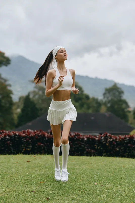 Young athletic woman jogging in a white sports outfit on a green field, showcasing vitality and energy in a natural setting