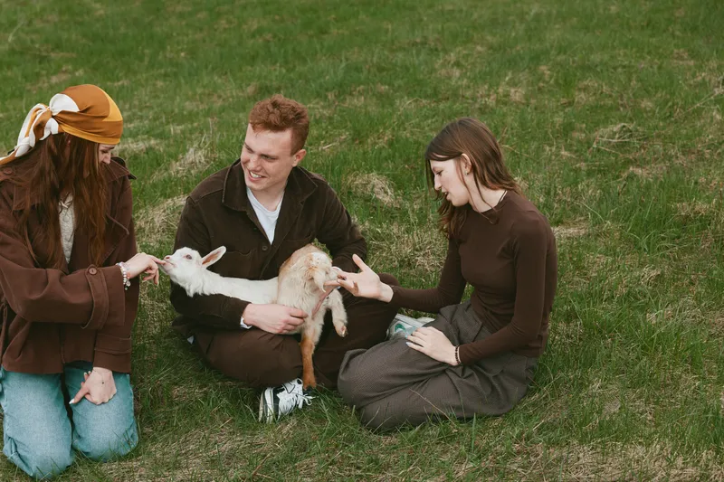 Lifestyle scene with two women and a man sitting on grass, gently feeding a young goat kid. Natural light creates a warm, relaxed mood of connection and rural friendship.