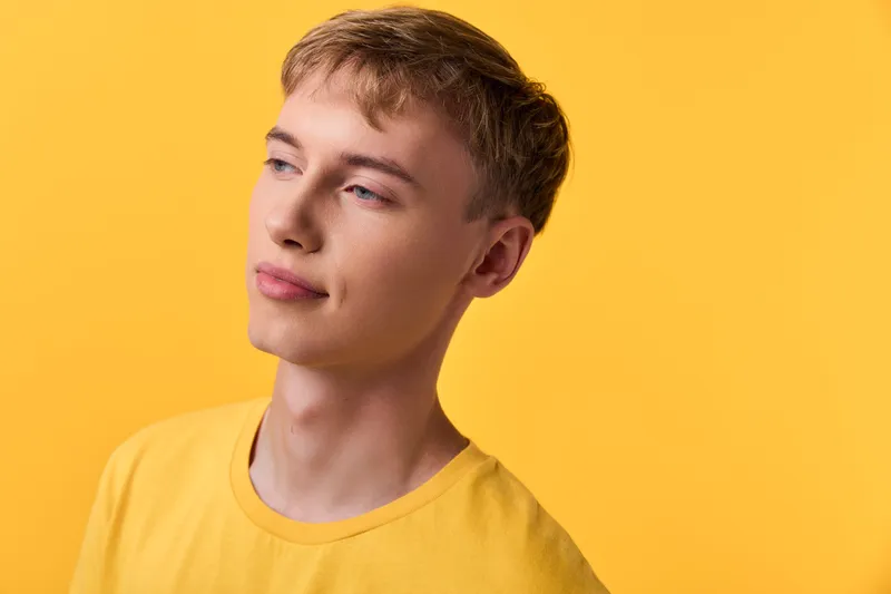 Young man in a yellow shirt gazing softly to the side against a vibrant yellow background, conveying calm, curiosity and casual style in a studio setting with warm light and minimal distractions