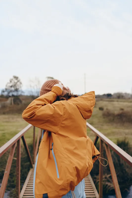 Woman lifestyle in outdoor countryside, wearing yellow coat and beanie, walking on a wooden bridge, hood up and wind, calm travel mood, autumn afternoon light