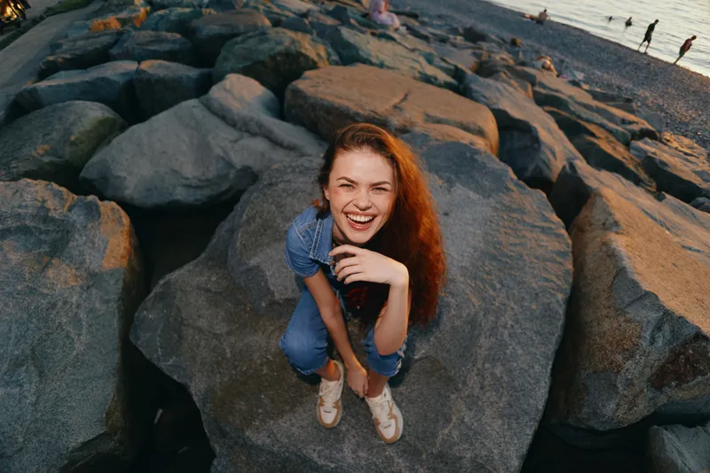 Lifestyle woman in modern boho-western denim street style laughing on rocky beach at sunset with film color tones enhancing natural mood and vibrant atmosphere