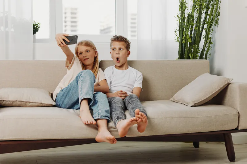 Two barefoot children sitting on beige sofa taking selfie with smartphone in bright living room. Casual home atmosphere and playful mood.