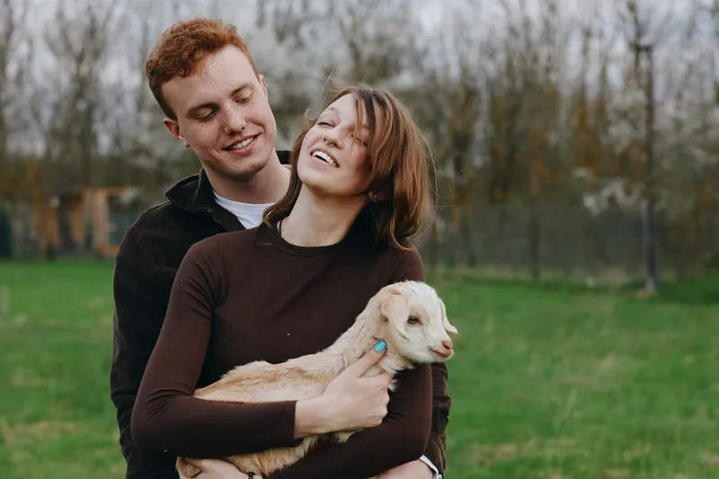Happy couple outdoors in a grassy field embrace a small goat kid, sharing warm affection and a joyful mood, capturing a rustic lifestyle with a friendly animal companion