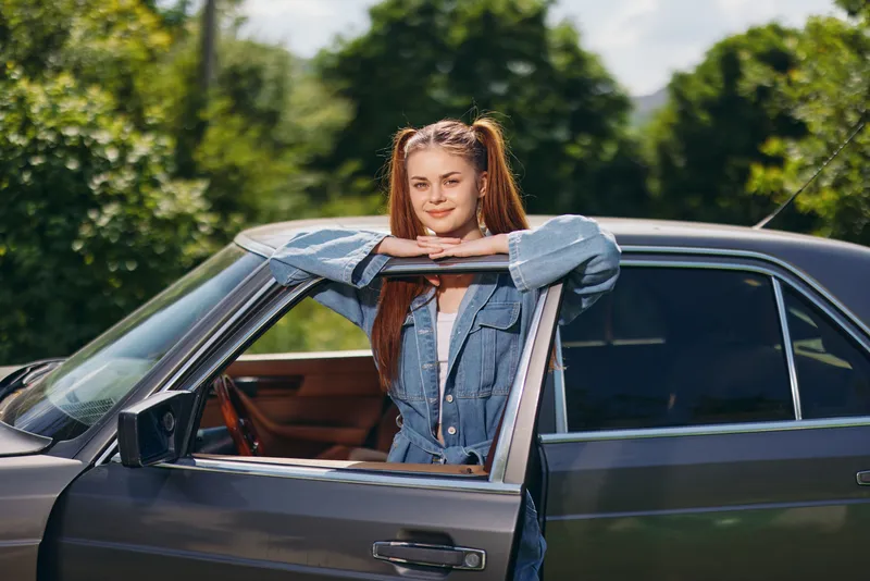 Young woman in casual denim outfit smiling by her vintage car, surrounded by greenery, reflecting a joyful summer vibe and freedom.
