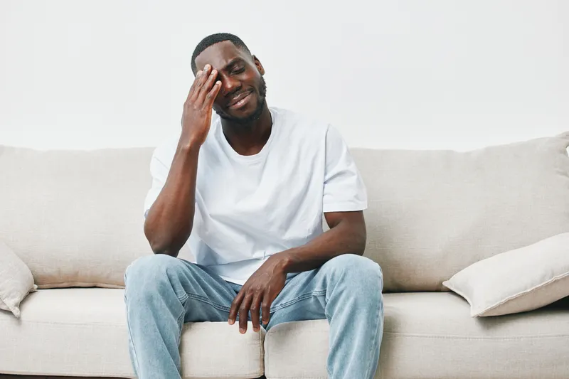 A young African American man sitting on a sofa, expressing a thoughtful emotion while holding his forehead, wearing a casual white t shirt and jeans