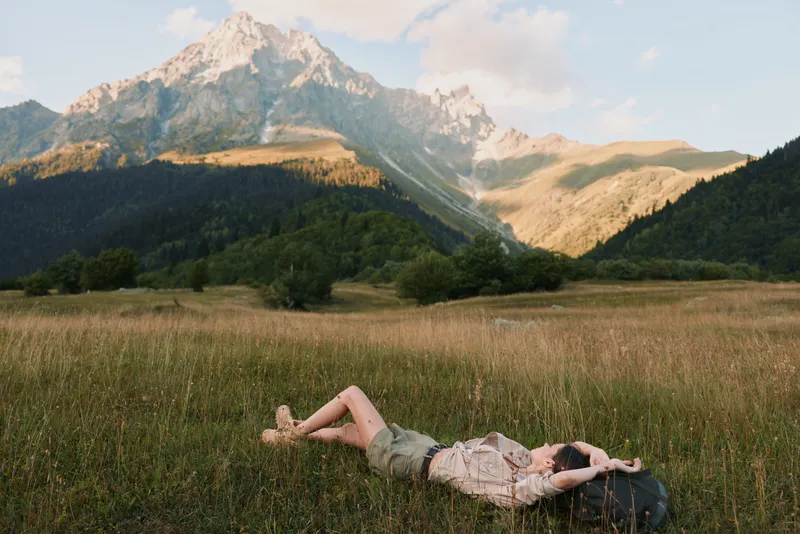Young woman lies on a grassy meadow with towering mountains in the background, enjoying a peaceful moment in nature during golden hour sunlight, casual dress and relaxed pose.