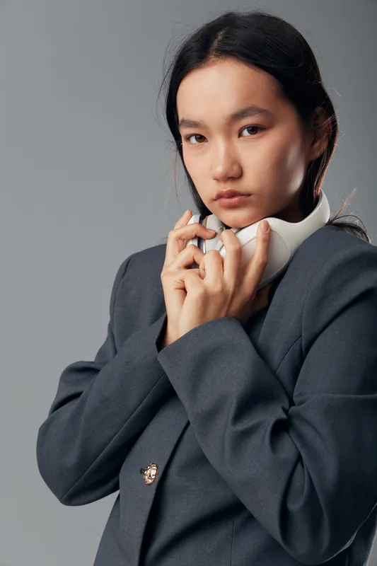 Young woman wearing headset and gray blazer standing with serious face on gray background. Asian businesswoman with dark hair and natural makeup holding headphones around neck.