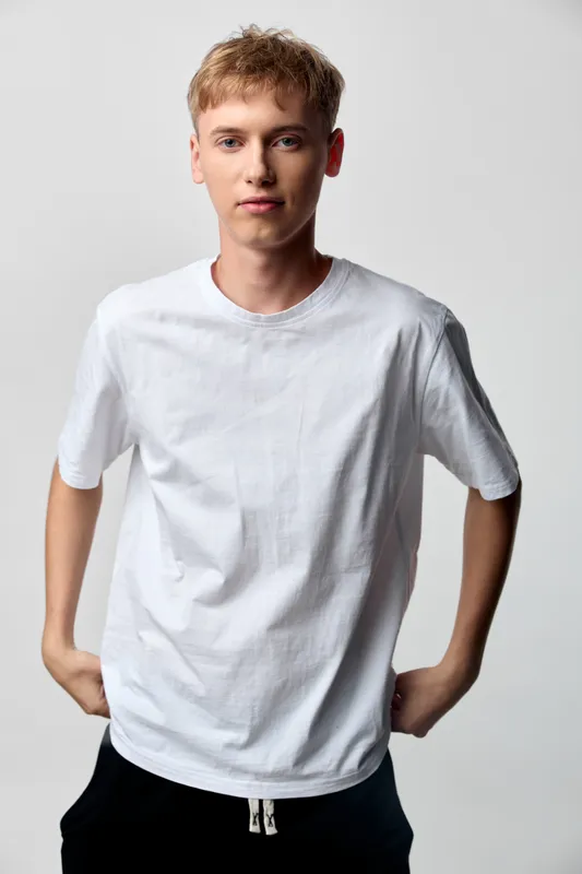 Young man in casual wear wearing a white tee, posed in a clean studio shot for fashion and model oriented lifestyle visuals, with a neutral backdrop, confident stance and minimalistic lighting.
