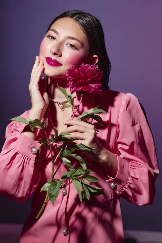 Young woman in pink elegant long sleeve dress holds bright red peony flower. She gently touches her face with manicured hand, makeup is vibrant and glossy. Studio lighting stylish concept.